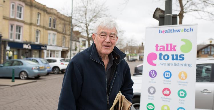 Elderly man standing in front of a Healthwatch banner saying "Talk to us"