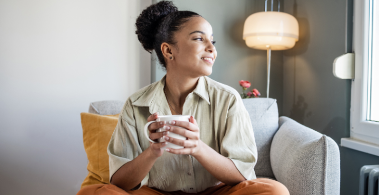 Woman sitting on a chair with her legs crossed and a mug in her hand as she looks at the window.