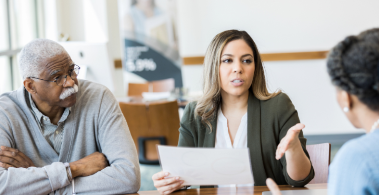 Two females and a male sitting around a table with one of the women holding a piece of paper and talking.