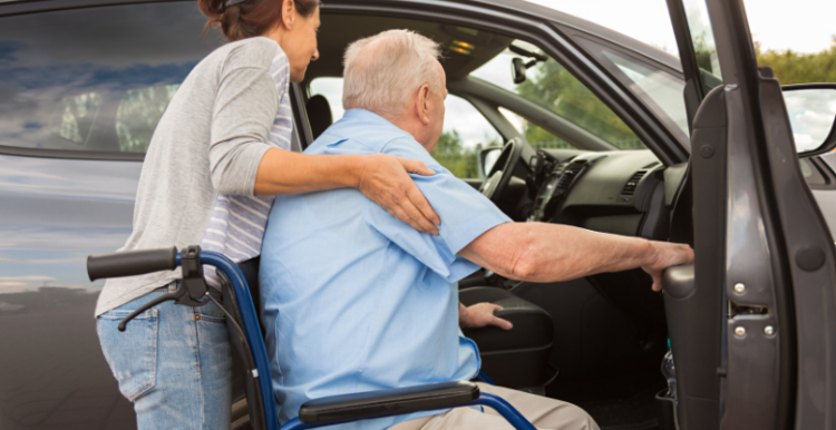 An elderly man sitting on a wheelchair being assisted in entering a car by a young lady.