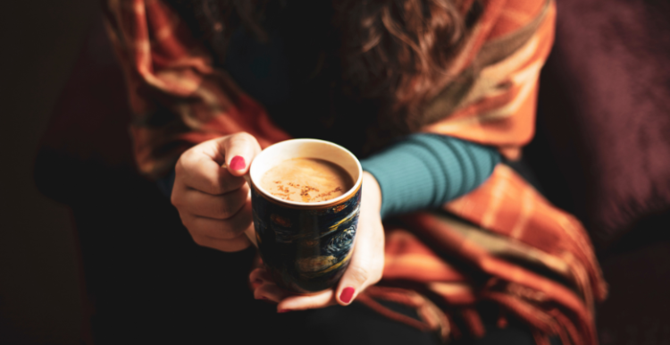 A woman keeping warm by holding a mug containing a hot drink and being wrapped in a blanket