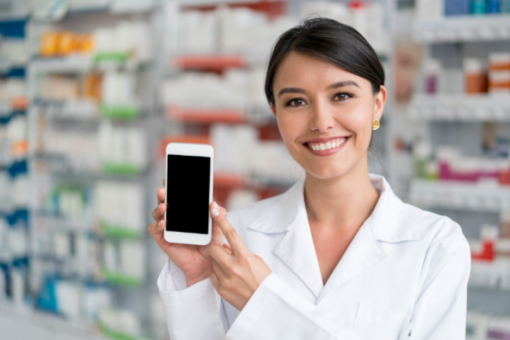 A female pharmacist smiling in a pharmacy and holding up a smartphone