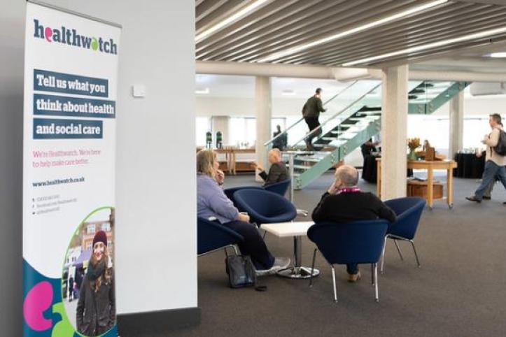 An open area inside a building with people sitting at tables and the Healthwatch banner.