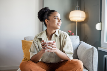 Woman sitting on a chair with her legs crossed and a mug in her hand as she looks at the window.