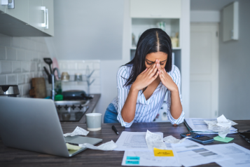 The image shows a woman in a kitchen looking stressed as she reviews papers and uses a laptop.