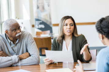Two females and a male sitting around a table with one of the women holding a piece of paper and talking.