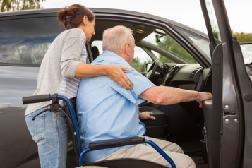 An elderly man sitting on a wheelchair being assisted in entering a car by a young lady.