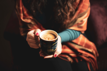 A woman keeping warm by holding a mug containing a hot drink and being wrapped in a blanket