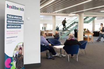 An open area inside a building with people sitting at tables and the Healthwatch banner.