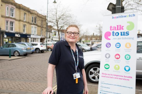 Female volunteer standing in front of a Healthwatch banner which says "Talk to us, we are listening"