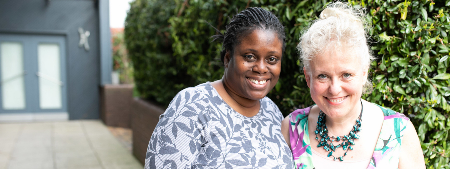 Two women standing in front of a hedge with their arms around each other and smiling at the camera